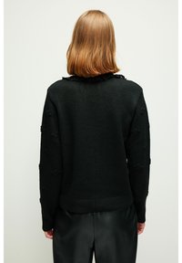 Svart stickad tröja med ett texturerat blommönster på ärmarna och ribbad nederkant. Har en volangkrage och knapprad vid axeln.