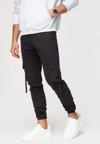 Homme portant un pantalon cargo noir avec des liens aux chevilles, des baskets blanches, un sweat-shirt gris et une montre-bracelet au poignet gauche.