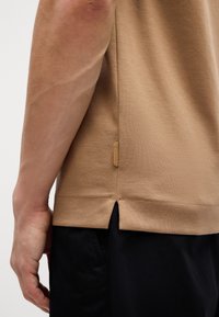 Beige bombažna majica s teksturirano površino, z bočnimi razporki in majhno rjavo oznako blizu roba, oblečena čez črne hlače.