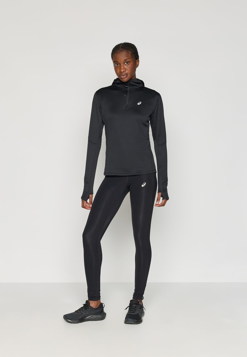 Zwarte sportieve top met lange mouwen en leggings van zacht, rekbaar materiaal, met een ritssluiting aan de voorkant, duimopeningen en een minimaal logo.