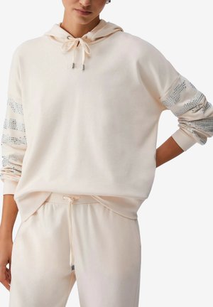 Felpa oversize beige chiaro con strisce di paillettes argento sulle maniche, abbinata a pantaloni con coulisse coordinati indossata da una persona con una mano sul fianco.