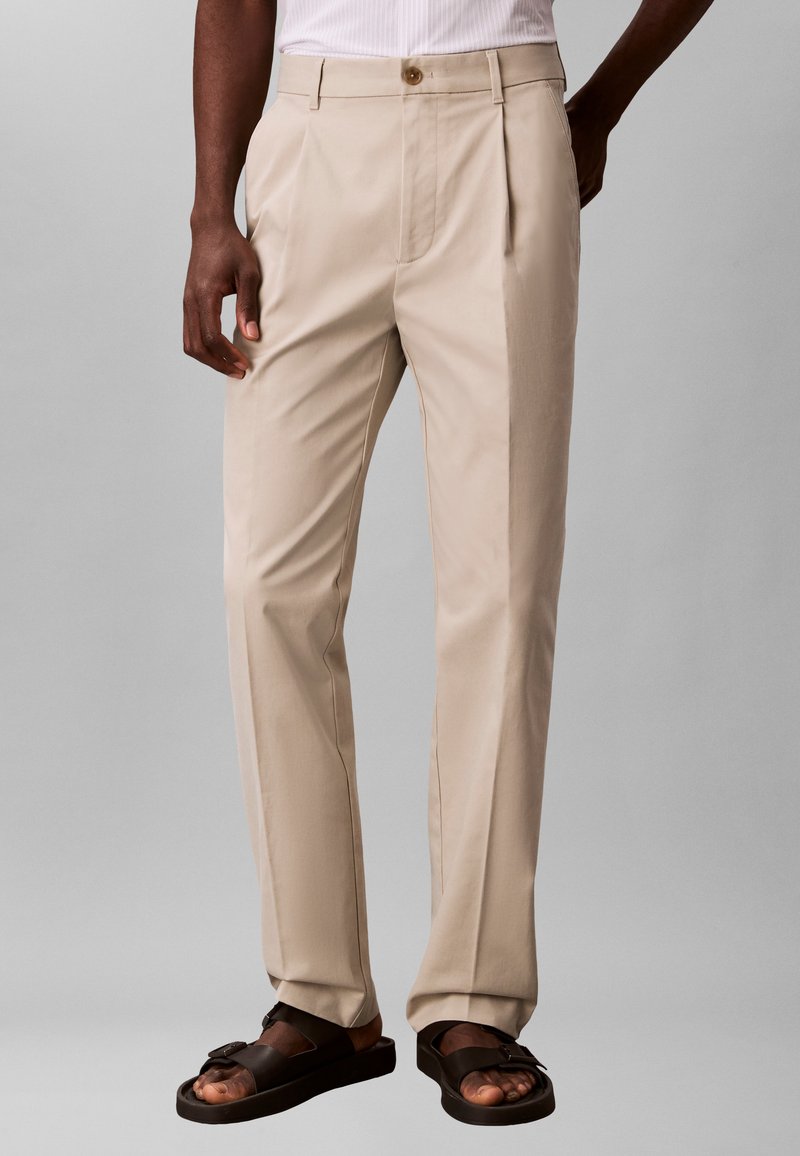 Homme portant un pantalon beige plissé et des sandales marron foncé, debout devant un fond gris uni.