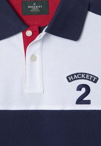 Polo en tissu texturé avec un schéma de couleurs bleu marine, blanc et rouge. Il dispose d'un col avec patte à deux boutons et d'un logo brodé avec le numéro "2".