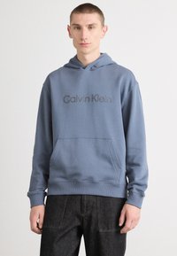 Zila kokvilnas kapuce ar priekšējo kabatu, ar pelēku logo "Calvin Klein" uz krūškurvja. Ribinātas aproces un apakša.