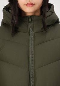 Chaqueta acolchada de color verde oliva con cierre frontal de cremallera y capucha. Presenta una textura suave y un patrón de acolchado en forma de chevrón para el aislamiento.