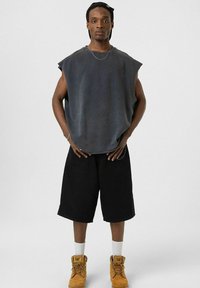 Grijze mouwloze kleding, ontspannen pasvorm, gecombineerd met zwarte shorts. Bruine werkschoenen, witte sokken, zilveren ketting en zwarte pet.