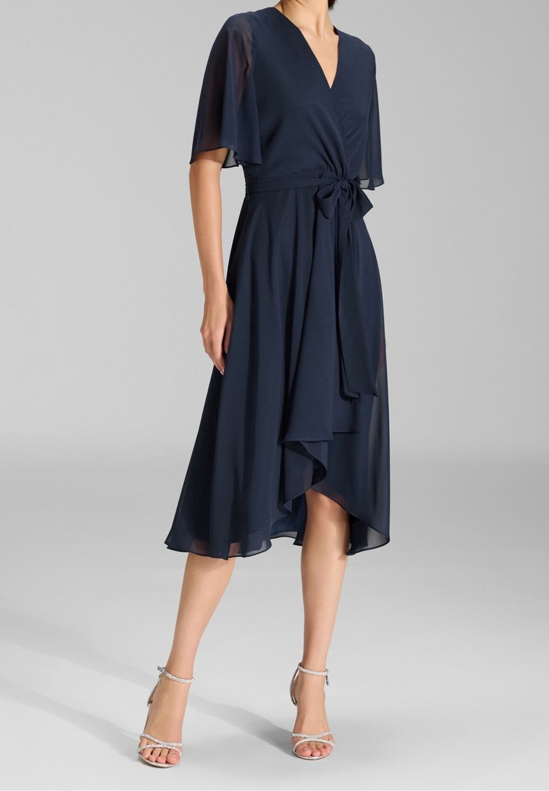 Femme portant une robe portefeuille bleu marine avec des manches volantées et un ourlet asymétrique, associée à des sandales à talons hauts argentées sur un fond gris.