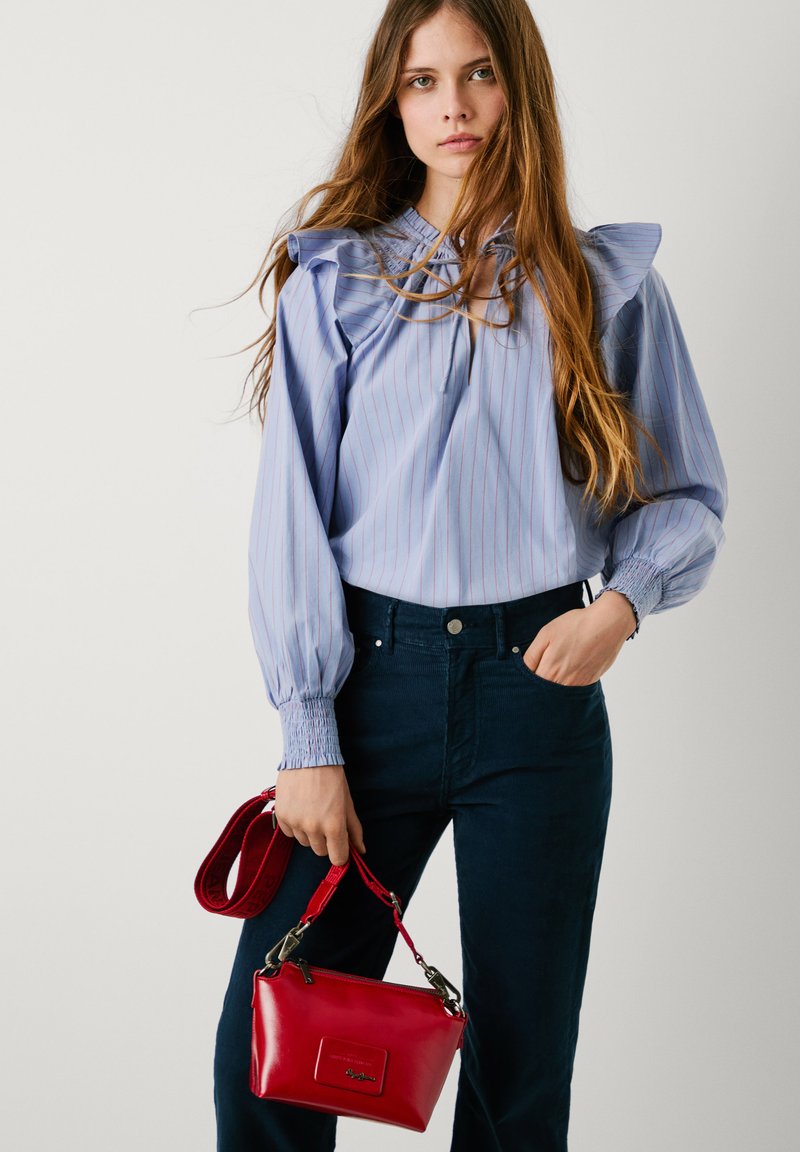 Frau mit langen Haaren, die eine blau gestreifte Bluse und schwarze Hosen trägt, hält eine kleine rote Handtasche mit einem dicken Trageriemen.