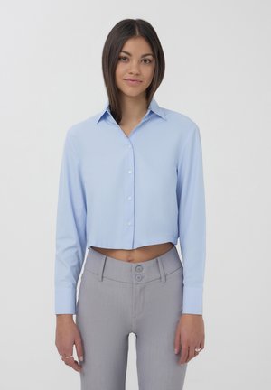 Camicia cropped azzurro chiaro con maniche lunghe e colletto, realizzata in tessuto morbido; abbinata a pantaloni grigi a vita alta con due bottoni.