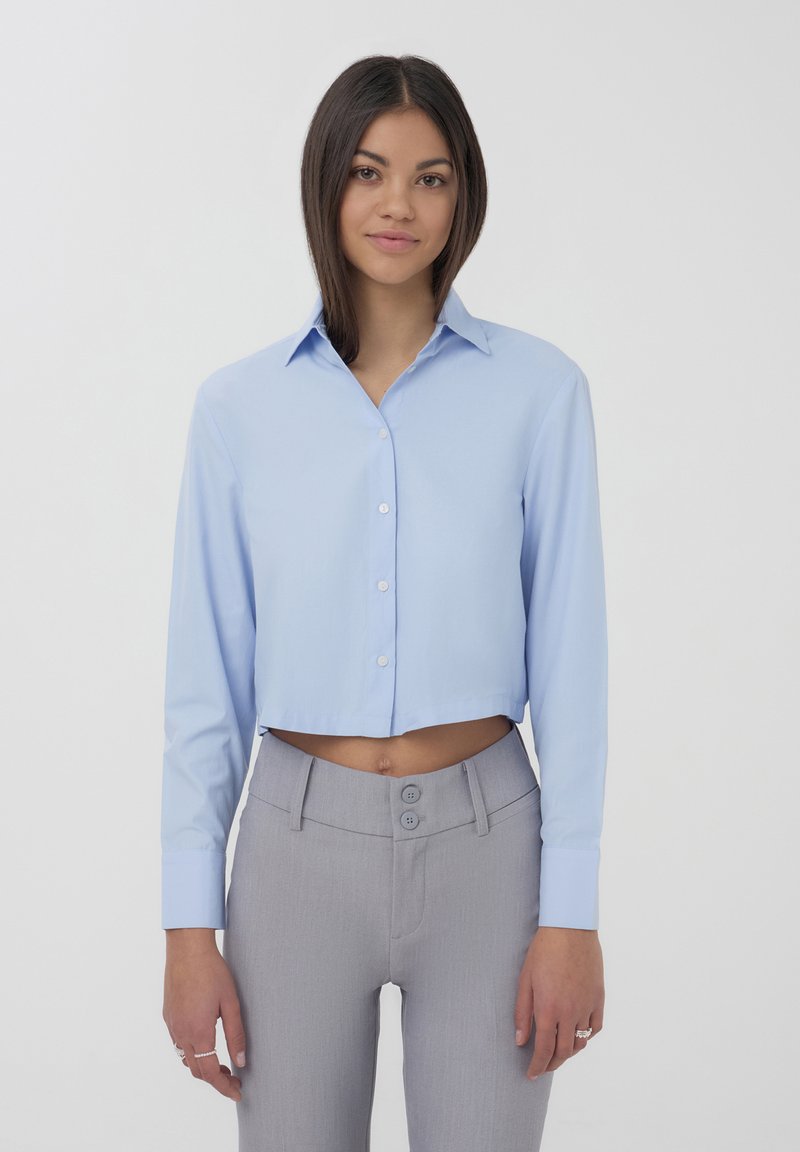 Camicia cropped azzurro chiaro con maniche lunghe e colletto, realizzata in tessuto morbido; abbinata a pantaloni grigi a vita alta con due bottoni.