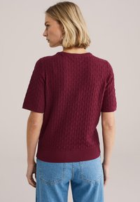 Kobieta z krótkimi blond włosami, ubrana w teksturowany bordowy sweter z krótkim rękawem i jasnoniebieskie dżinsy, zwrócona tyłem.