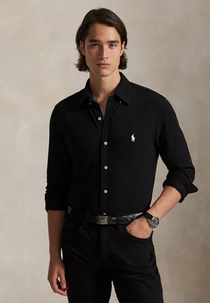 Un homme pose devant un fond beige uni. Il porte une chemise noire boutonnée et un pantalon, une montre, et a une main dans la poche.