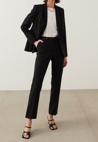 Costume noir ajusté avec un blazer cintré et un pantalon droit, associé à une chemise blanche et des sandales à talons strappy noires.