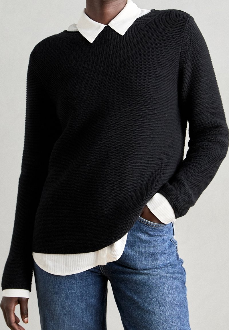 Personne portant un pull en maille noire par-dessus une chemise blanche à col, et un jean bleu, une main dans la poche, sur un fond uni.