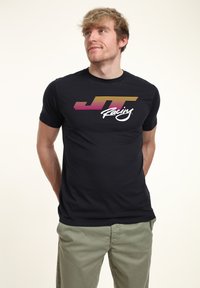Svart kortärmad t-shirt med en flerfärgad "JT Racing"-logotyp med horisontella ränder i rosa, gult och vitt. Tillverkad av bomull.