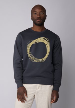 Navy sweatshirt made of soft fabric featuring a large, light yellow circular design. Long sleeves and ribbed cuffs. Worn with light-colored pants.