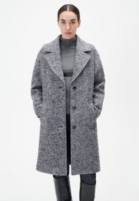 Grau und schwarz gestreifter Mantel im Fischgrätenmuster mit großen Revers, Frontknöpfen und Seitentaschen, getragen über einem grauen Rollkragenpullover und schwarzen Hosen.