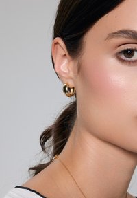 Złote kolczyki w kształcie obręczy z kręconym wzorem, o gładkim metalicznym wykończeniu i lekkiej powierzchni odblaskowej. Noszone na uchu modelki.