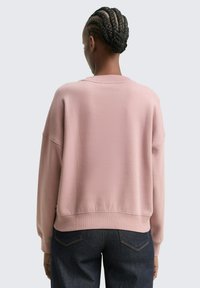 Persona con un moño trenzado que lleva una sudadera ligera de color rosa claro y jeans azul oscuro, de espaldas frente a un fondo liso.