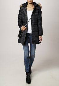 Abrigo de plumas negro con capucha de pelo, cierre con cremallera y puños acanalados, combinado con una camiseta blanca y vaqueros azules, llevado con botas negras.