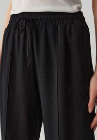 Zwarte, lichte broeken met een elastische tailleband en trekkoord. Beschikt over een gladde textuur en zijplooien voor een relaxte pasvorm.