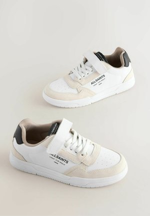 Bílé a béžové tenisky s kombinací hladké kůže a semiše, perforovanou špičkou, nastavitelným páskem a černými akcenty na patě.