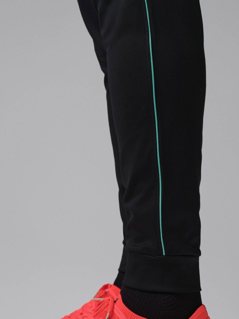 Onderbeen met zwarte broek met een dunne turkooizen zijstreep en felrode sportschoenen tegen een grijze achtergrond.