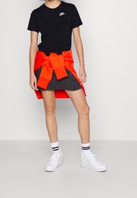 Person som bär en svart Nike-t-shirt, svart randig kjol, vita sneakers med randiga strumpor, samt en rödaktig-orange sweatshirt knuten runt midjan.