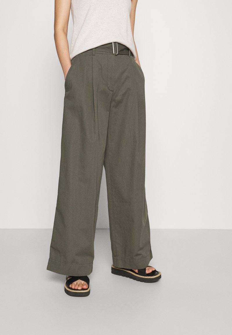 Wide-leg trousers in olive green cotton, featuring front pleats, side pockets, and a smooth belt with a metal buckle. Paired with black sandals.