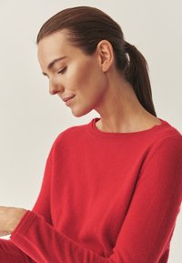 Sarkanais vilnas džemperis ar apaļu kakla izgriezumu un garām piedurknēm, ar mīkstu tekstūru un ribotiem aprocēm. Modelis ir nedaudz pagriezies un iesaistīts aktivitātē.