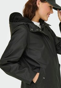 Frau in einer schwarzen, gefütterten, wasserdichten Jacke mit Kapuze, darunter ein weißes Hemd, während sie eine schwarze Baseballkappe eingestellt.
