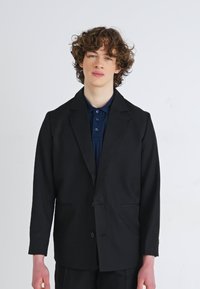 Giacca blazer nera con una vestibilità su misura, chiusura con un bottone singolo e rever a punta. Indossata sopra una polo blu scuro.