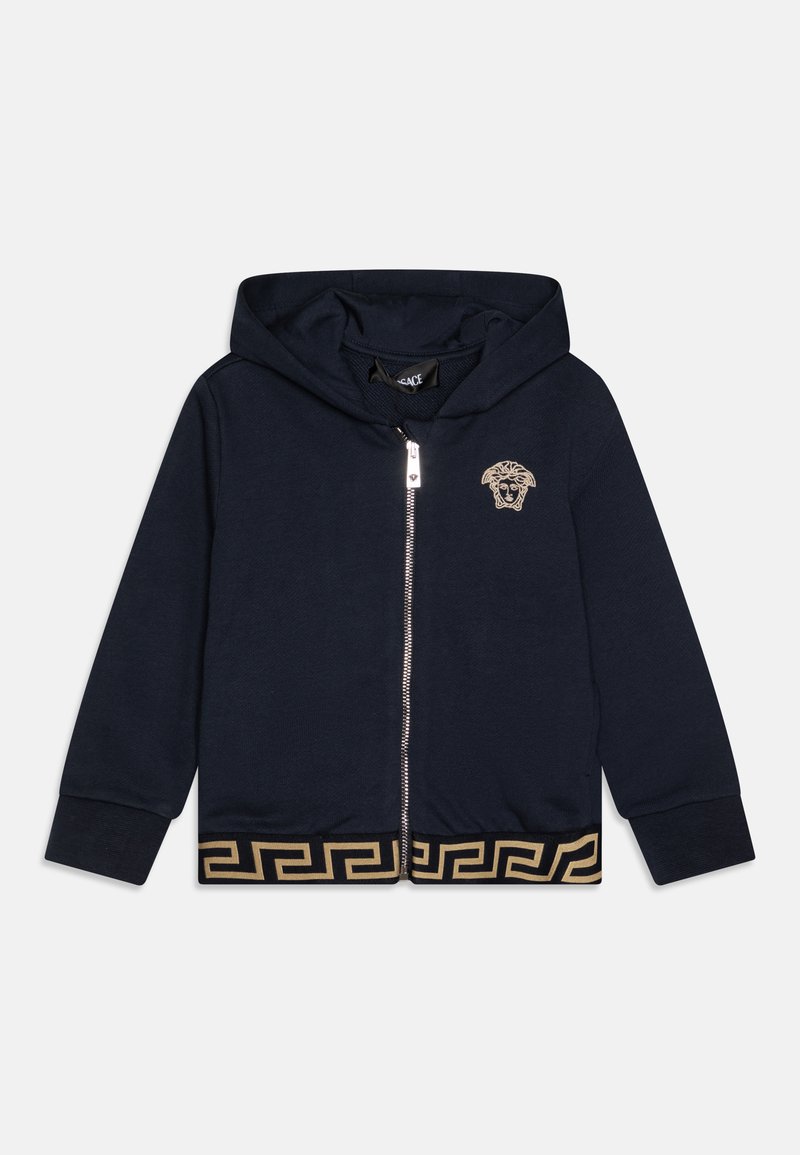 Sudadera con cremallera en color navy, hecha de una tela suave, que presenta un logo dorado de cabeza de león en el pecho y un patrón de llaves griegas en dorado en el dobladillo.