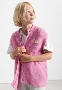 Roza rebrasta pletena brezrokavnika s kratkimi rokavi, sprednjimi gumbi in majhno rumeno cvetno nalepko. Nosi se preko bele majice, kar poudarja mehko teksturo.