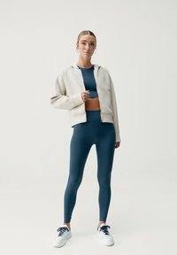 Felpa beige con zip sopra un top sportivo blu a maniche corte e leggings a vita alta coordinati, completata da sneakers bianche con accenti blu navy.