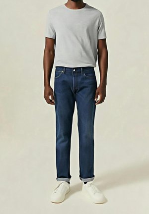 Homme portant un t-shirt gris clair uni à manches courtes, un jean bleu foncé retroussé, et des baskets blanches, debout devant un fond neutre.