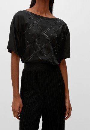 Kvinde iført en sort kortærmet top med skinnende diagonale linjer og sorte teksturerede bukser med høj talje mod en ensfarvet baggrund.