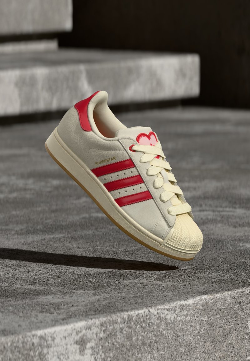 Baskets beiges avec trois bandes rouges, embout en caoutchouc, design à lacets et un accent rouge au talon. Marque "Superstar" sur le côté.