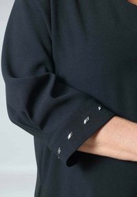 Zwarte stoffen top met een textuurmouwmanchet versierd met zilveren vierkante accenten, die de ontspannen uitstraling en de gladde textuur benadrukt.