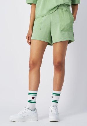 Grüne Baumwollshorts mit lockerer Passform, ausgestattet mit Seitentaschen. Kombiniert mit weißen Sneakern und weißen Socken mit grünen Streifen.