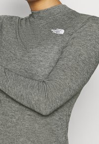 Šedý dlouhý rukáv top s vysokým výstřihem, vyrobený z texturované látky. Obsahuje malé bílé logo na hrudi značky The North Face.