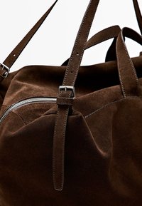 Brun mockaväska med dragkedja, utrustad med två justerbara remmar och silverfärgade detaljer, som visar upp mjuk textur och enkel design.