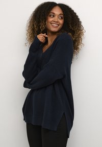 Tumesinine ülemahutav kampsun soonilise tekstuuri, V-kaeluse ja külgmistest lõhikestega; kombineeritud mustade pükstega.