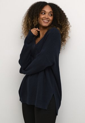 Granatowy, oversize'owy sweter z prążkowanej tkaniny, z dekoltem w serek i bocznymi rozcięciami. Modelka pozuje z założonymi rękami, podkreślając luźny krój.