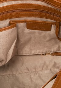 Béžový interiér kabelky Michael Kors s vzorem loga, hnědé kožené obložení, více kapes včetně zipové kapsy a otevřených přihrádek.