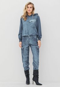 Moteris vilkinti mėlynai pilką gobtuvą su kišene, suderintomis kargo kelnėmis ir juodais siaurais aukštakulniais bateliais, stovi prieš neutralų foną.