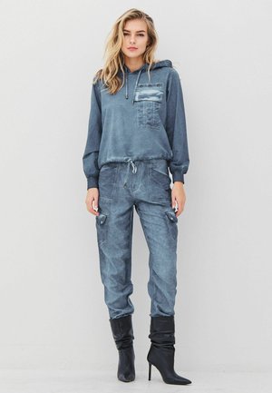 Vrouw in een blauw-grijze hoodie met een borstzak, bijpassende cargobroek en zwarte spitse hoge hakken, staande tegen een effen achtergrond.