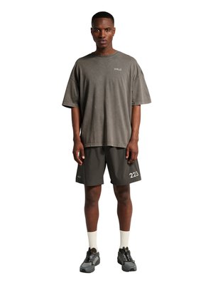 Mann steht mit dem Gesicht nach vorne und trägt ein übergroßes graues T-Shirt mit dem Schriftzug "HALO", dunkle Shorts mit den Logos "223" und "HALO", weiße Socken und schwarze Sneakers.