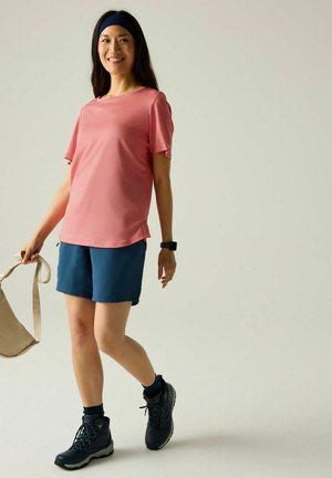 Kobieta ubrana w różowy T-shirt z krótkim rękawem, niebieskie szorty, buty trekkingowe i opaskę na głowę, uśmiechająca się trzymająca beżową torbę.