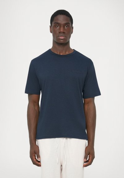 Hombre vestido con una camiseta de manga corta azul marino lisa y pantalones de tiro con cordón de color claro, de pie frente a un fondo blanco.